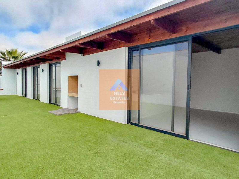 Spacious outdoor patio area featuring artificial lawn, exposed timber rafters, and a built-in braai, seamlessly connected to the interior via large sliding glass doors.