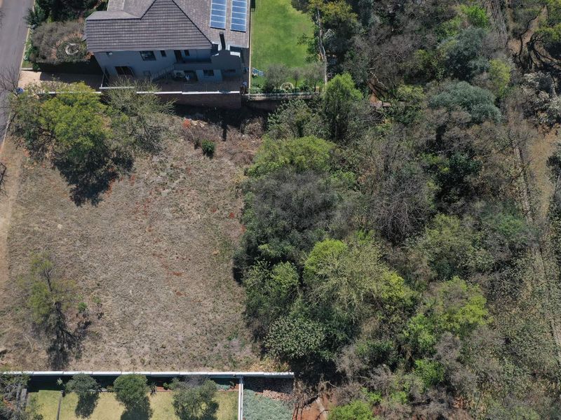 Aerial view of a vacant residential plot featuring established trees and direct street access, situated adjacent to existing residential properties.