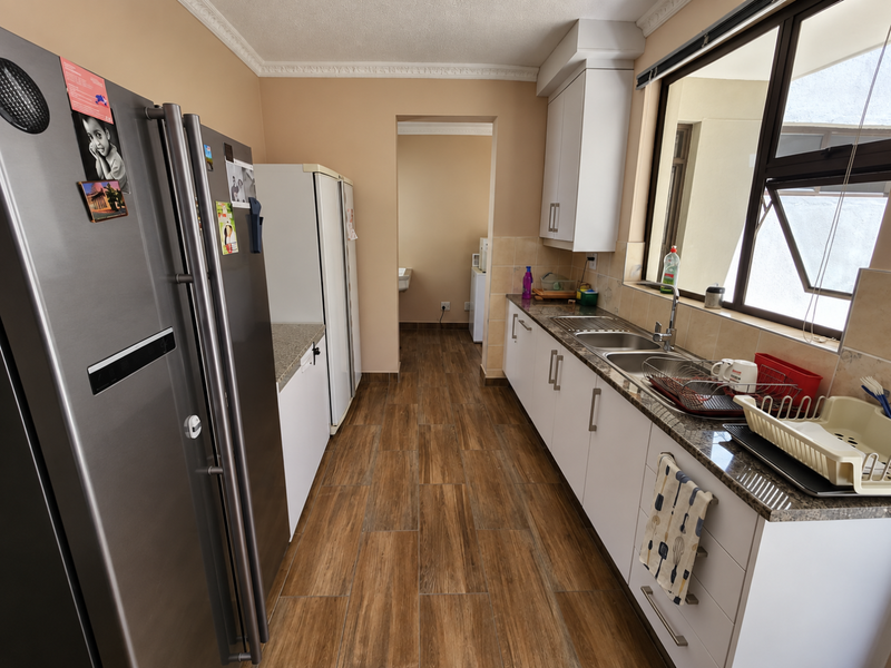 A functional kitchen featuring granite countertops, white cabinetry, a stainless steel double sink, and wood-look tiled flooring with a connecting scullery area.