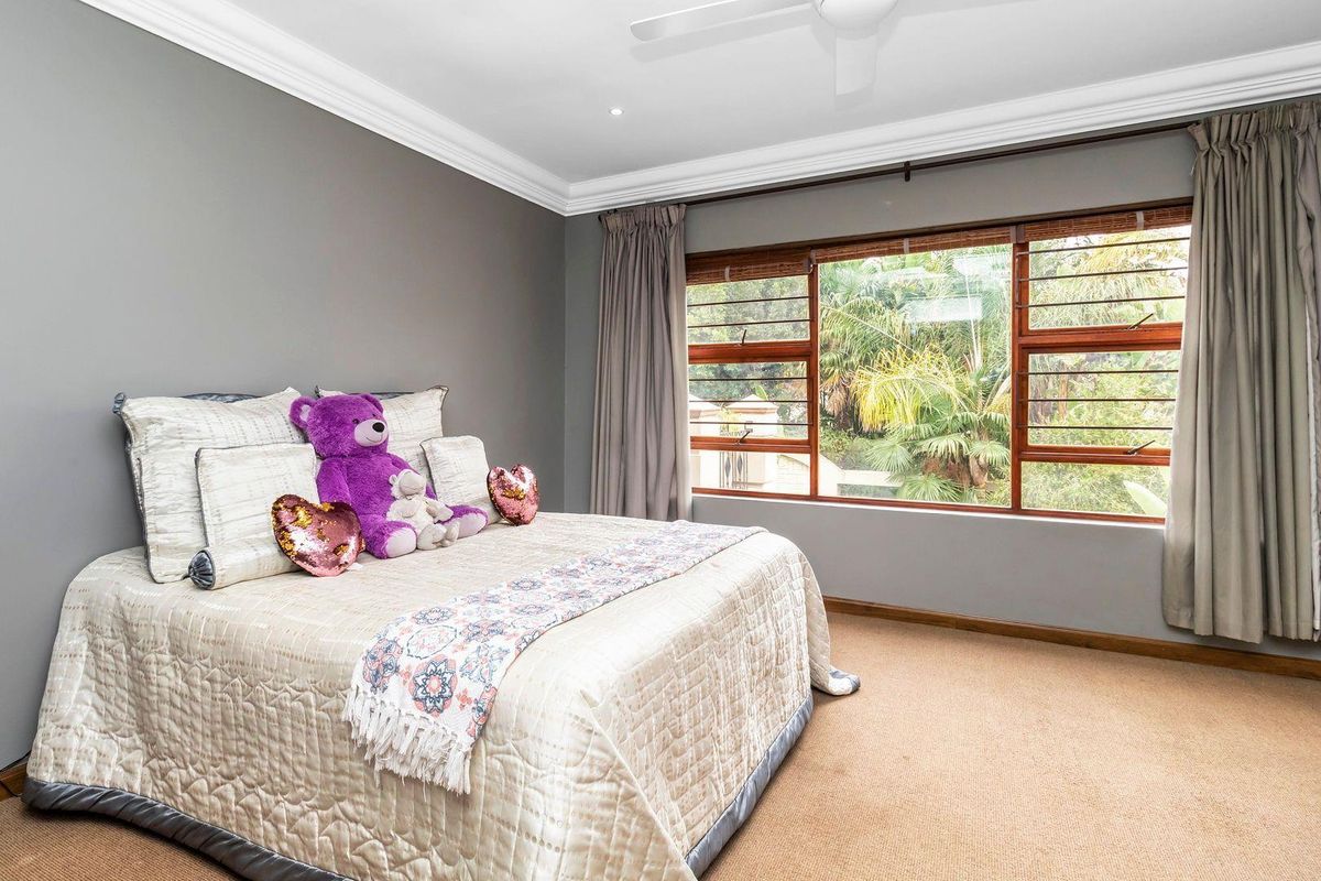 A well-maintained bedroom featuring neutral grey walls, plush carpeting, crown molding, and large wooden-framed windows overlooking lush greenery.
