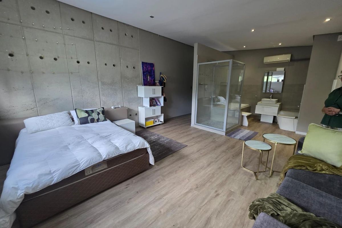 Spacious open-plan bedroom suite featuring a contemporary concrete feature wall, integrated en-suite bathroom with glass shower enclosure, and modern wood-look flooring.