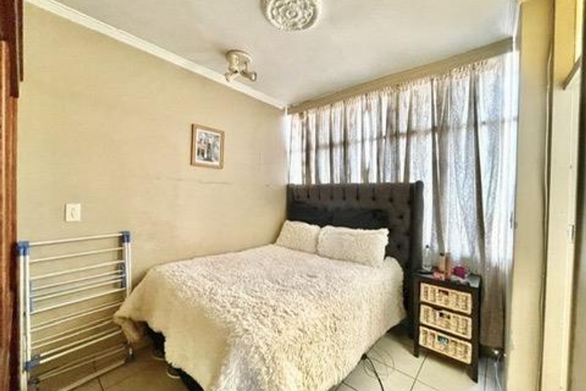 A bedroom featuring tiled flooring, a ceiling rose, and large windows with curtains, providing a functional living space.