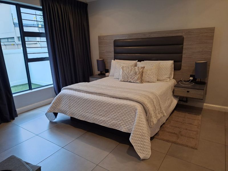 A well-maintained bedroom featuring tiled flooring, a modern built-in headboard with integrated floating bedside tables, and large windows with security bars.