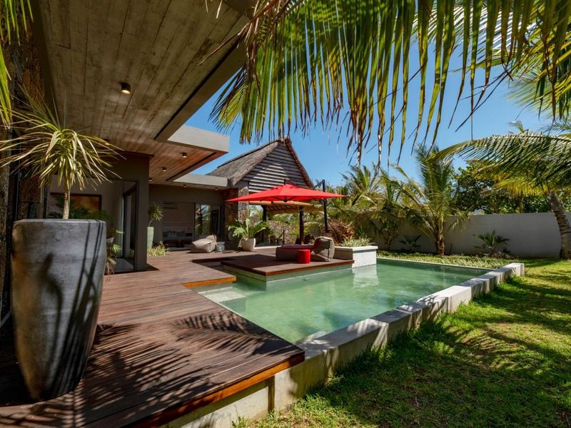 A beautifully landscaped outdoor pool area featuring a modern timber deck, concrete pool surround, and a covered patio area.