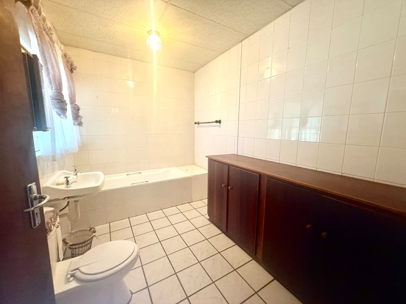 A dated bathroom featuring a bathtub, pedestal basin, toilet, and extensive built-in wooden cabinetry with white tiled walls and flooring.