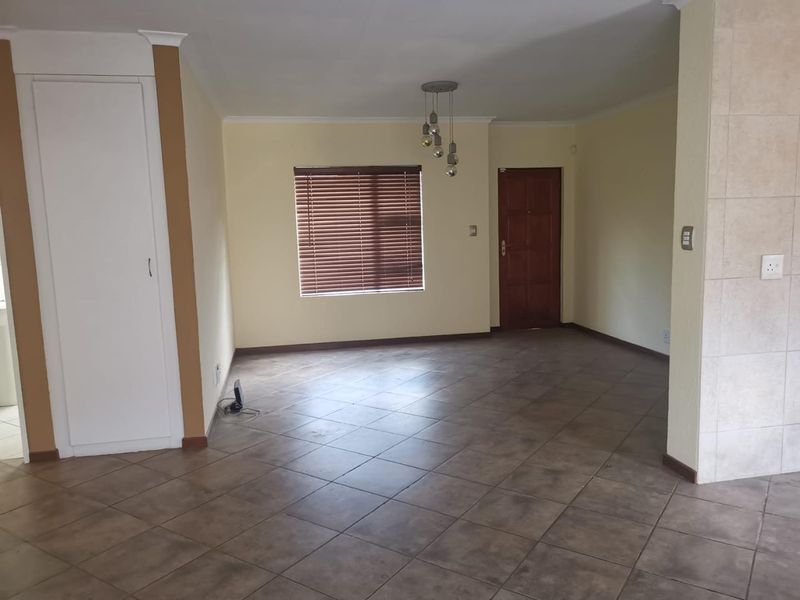 A spacious, empty living area featuring neutral walls, tiled flooring, a built-in cupboard, and a solid wood entrance door with venetian blinds.