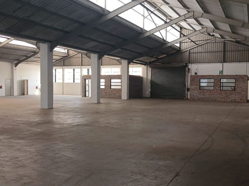 Spacious industrial warehouse featuring high ceilings with steel trusses, concrete flooring, a roller shutter door, and an integrated brick office partition.