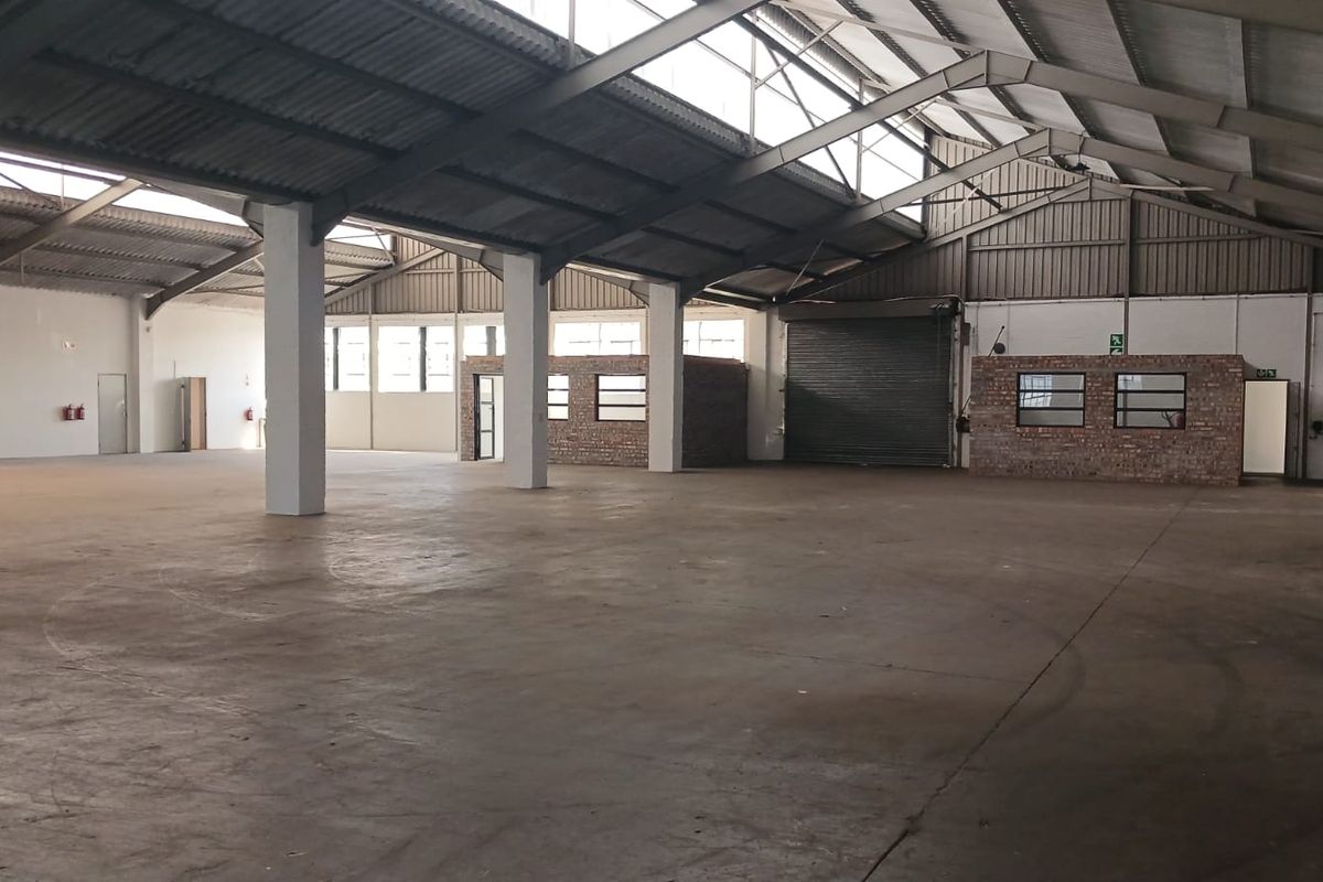 Spacious industrial warehouse featuring high ceilings with steel trusses, concrete flooring, a roller shutter door, and an integrated brick office partition.