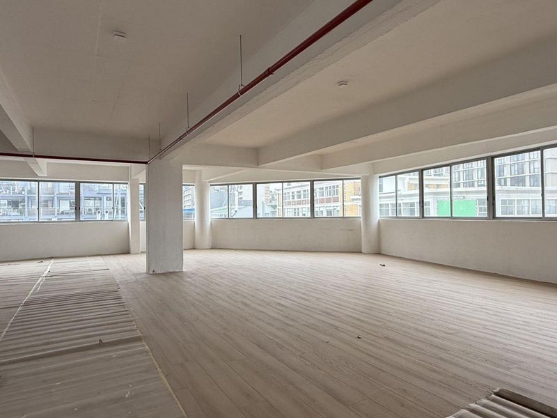 A spacious, open-plan office area featuring large windows, neutral laminate flooring, and exposed fire sprinkler piping, offering a bright and versatile workspace.