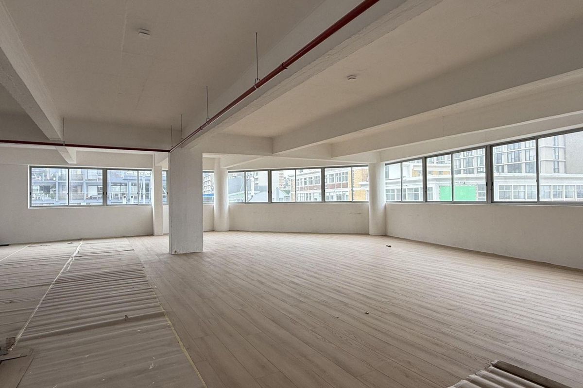 A spacious, open-plan office area featuring large windows, neutral laminate flooring, and exposed fire sprinkler piping, offering a bright and versatile workspace.