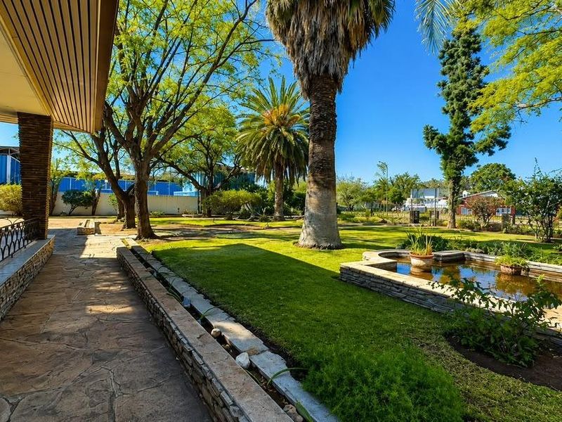 A well-maintained garden featuring a stone-paved walkway, a covered patio area with stone pillars, and a decorative pond set within a landscaped lawn.
