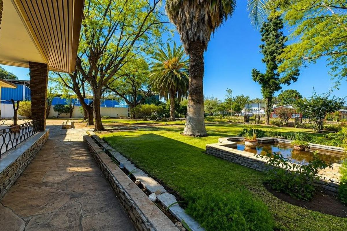 A well-maintained garden featuring a stone-paved walkway, a covered patio area with stone pillars, and a decorative pond set within a landscaped lawn.