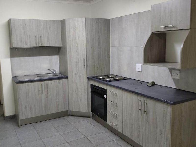 A functional kitchen featuring light wood-grain cabinetry, dark laminate countertops, a built-in oven with hob, and neutral tiled flooring.