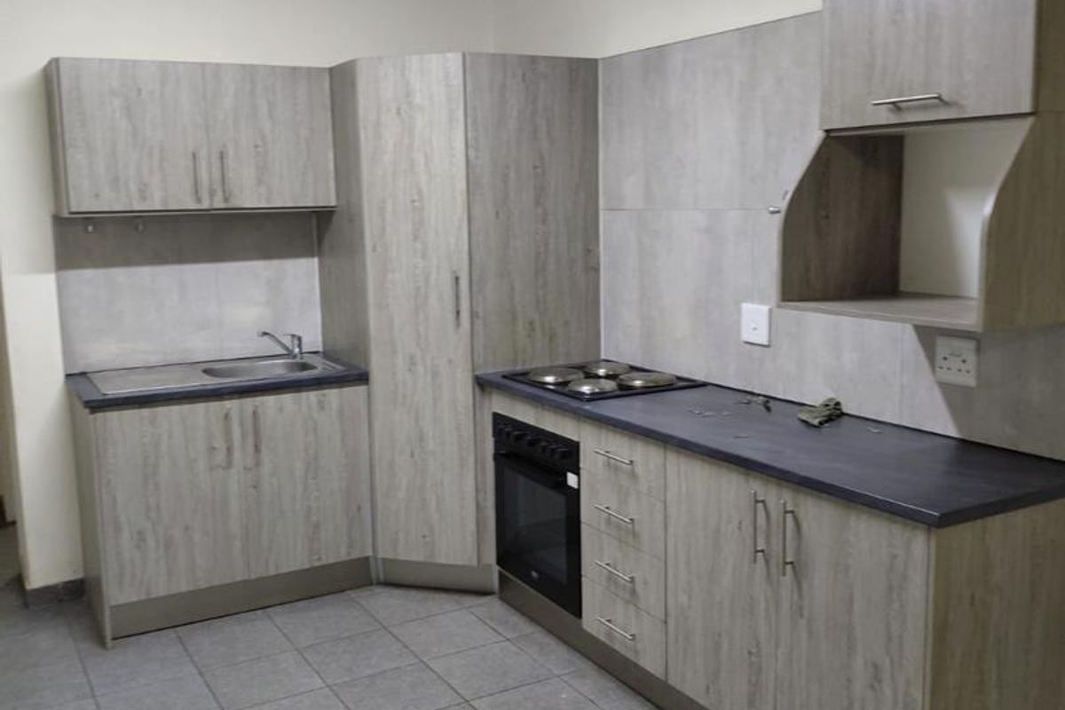 A functional kitchen featuring light wood-grain cabinetry, dark laminate countertops, a built-in oven with hob, and neutral tiled flooring.