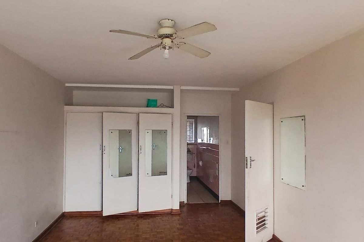 A vacant bedroom featuring parquet flooring, built-in cupboards with mirrored panels, and a ceiling fan, with direct access to an en-suite bathroom.