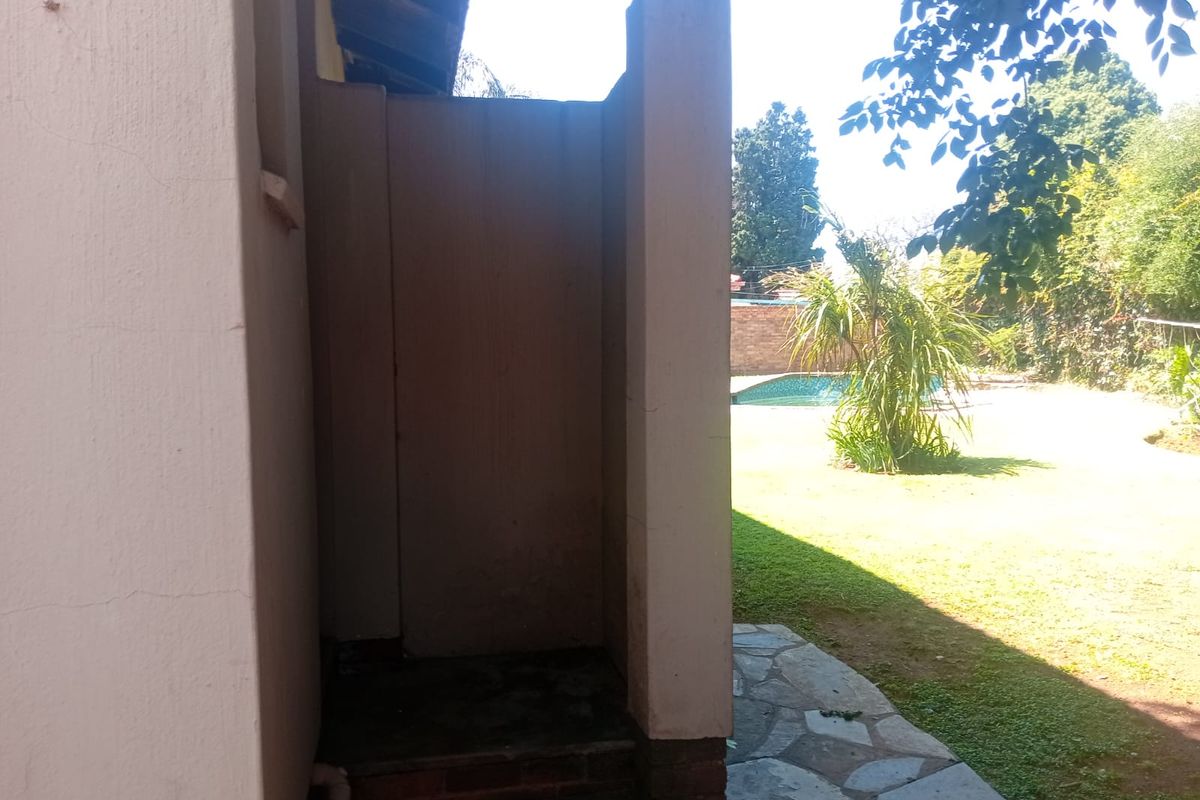 Rear exterior view featuring an outdoor shower enclosure, stone paved pathway, and a view of the garden and swimming pool area.