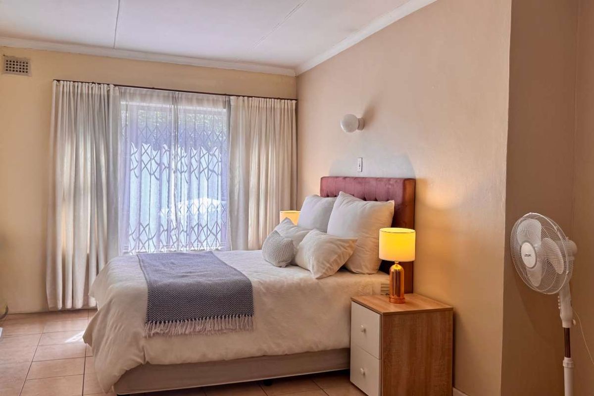 A bedroom featuring tiled flooring, a built-in headboard, and large windows with security gates, providing a functional and comfortable sleeping space.