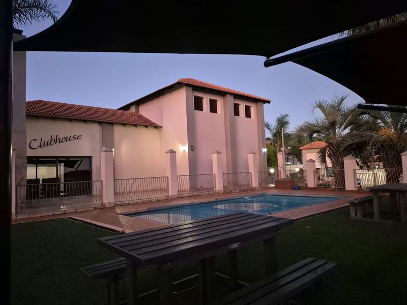 Communal pool area featuring a swimming pool, paved surround, and adjacent clubhouse building with tiled roofing.