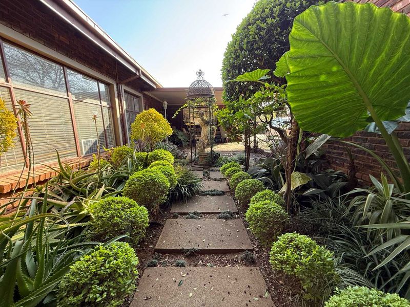 A well-maintained garden path featuring stone pavers, lush manicured shrubbery, and mature greenery leading toward a covered patio area.