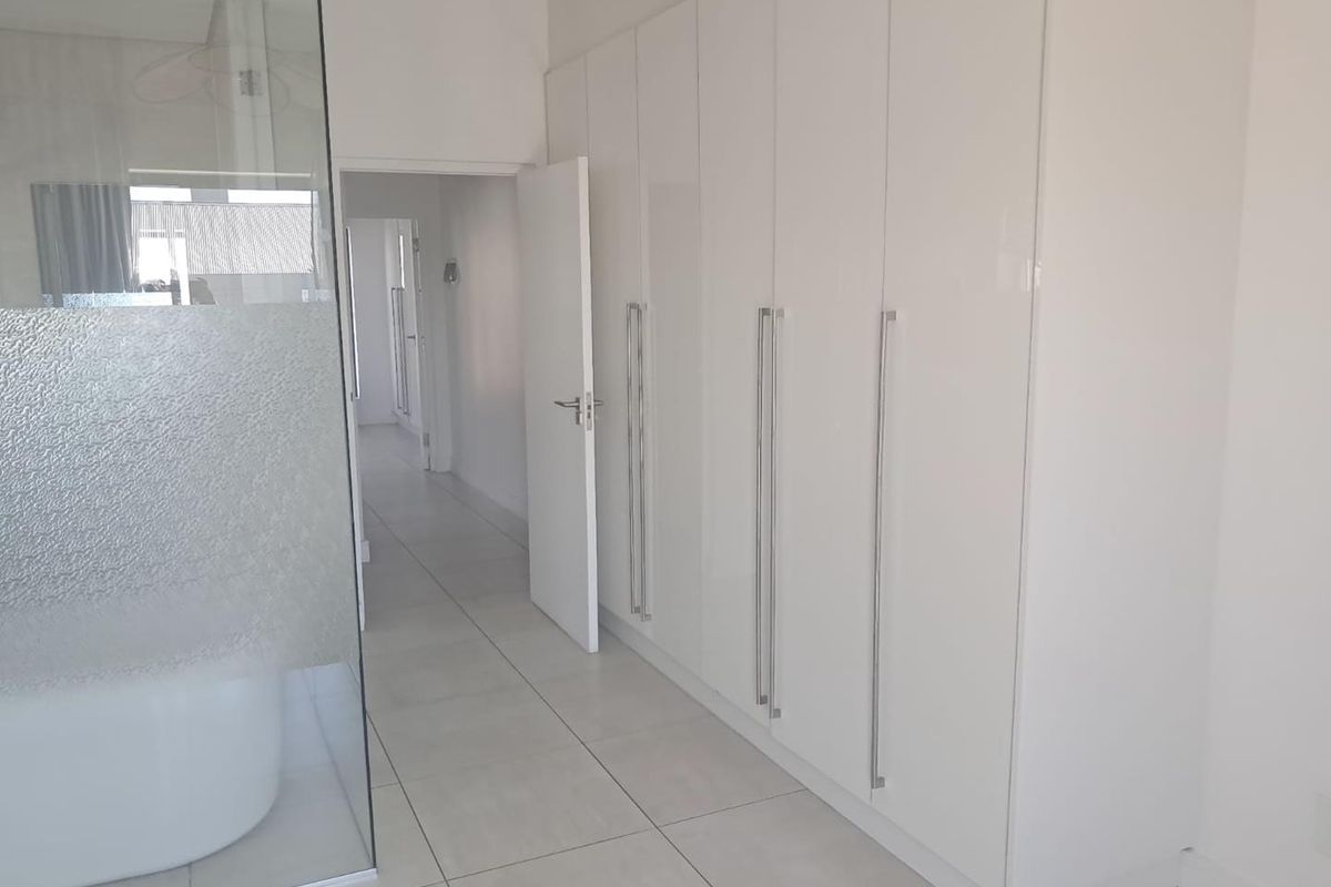 A bright bedroom featuring extensive built-in cupboards, sleek tiled flooring, and a modern glass partition, leading into a hallway.