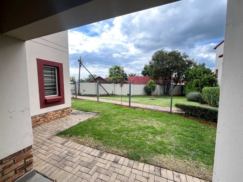 A private rear garden featuring a paved patio area, well-maintained lawn, and secure wire mesh fencing, viewed from under a covered patio.