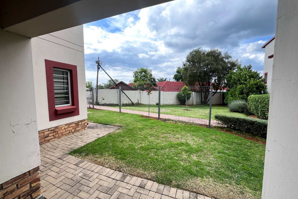 A private rear garden featuring a paved patio area, well-maintained lawn, and secure wire mesh fencing, viewed from under a covered patio.