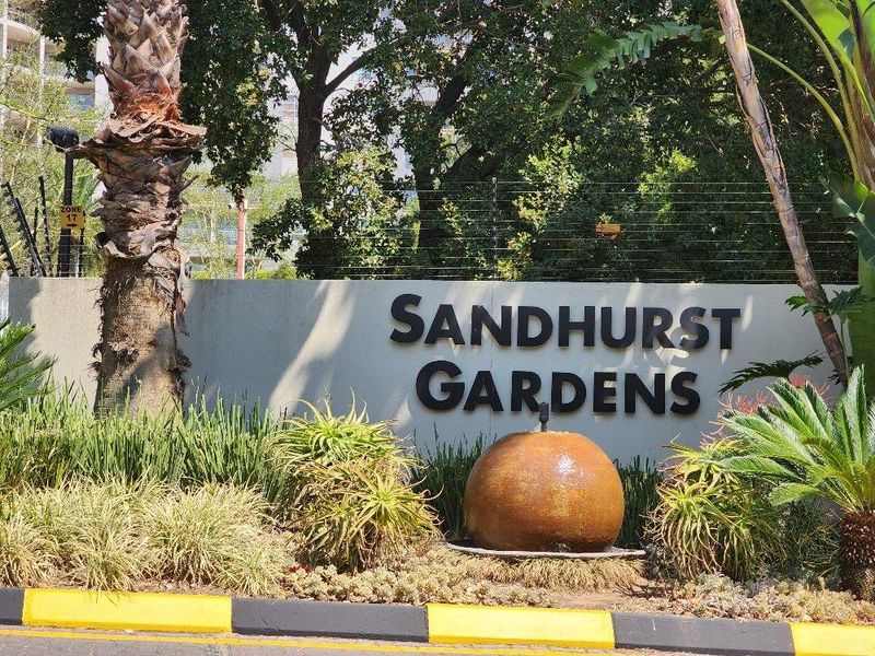 The entrance signage and landscaped garden area of Sandhurst Gardens, featuring a concrete boundary wall, electric fencing, and a decorative water feature.