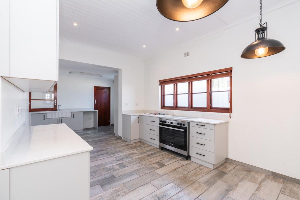 A bright, modern kitchen featuring light grey cabinetry, stone countertops, a farmhouse sink, and a freestanding oven, with neutral tiled flooring throughout.