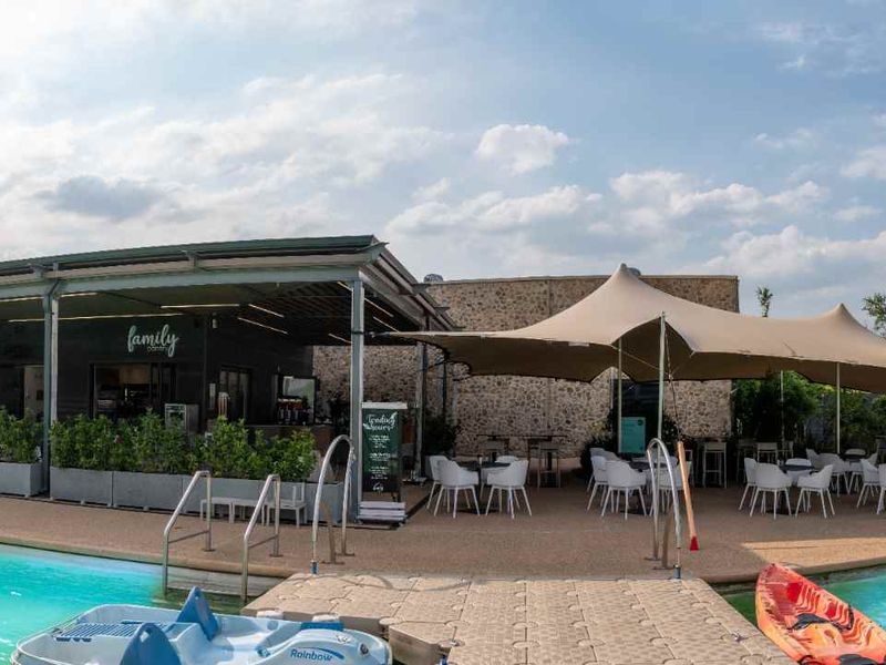 A spacious outdoor pool area featuring a large swimming pool, a covered patio with shade sails, and stone-clad building exterior.