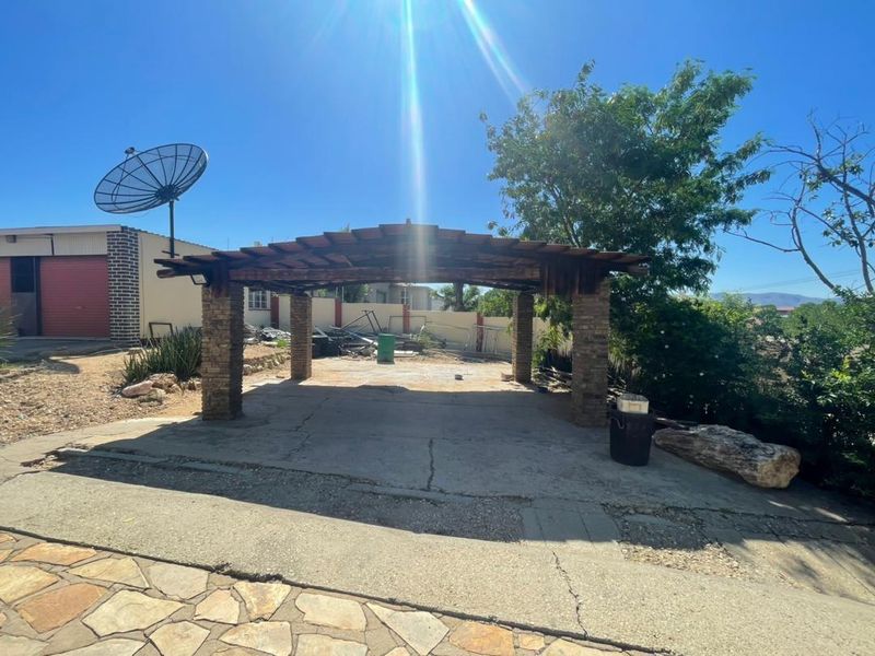 An outdoor covered patio area featuring stone-clad pillars and a timber pergola, set within a spacious yard with a concrete base.