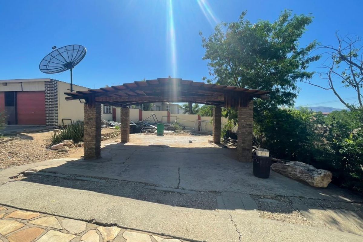 An outdoor covered patio area featuring stone-clad pillars and a timber pergola, set within a spacious yard with a concrete base.