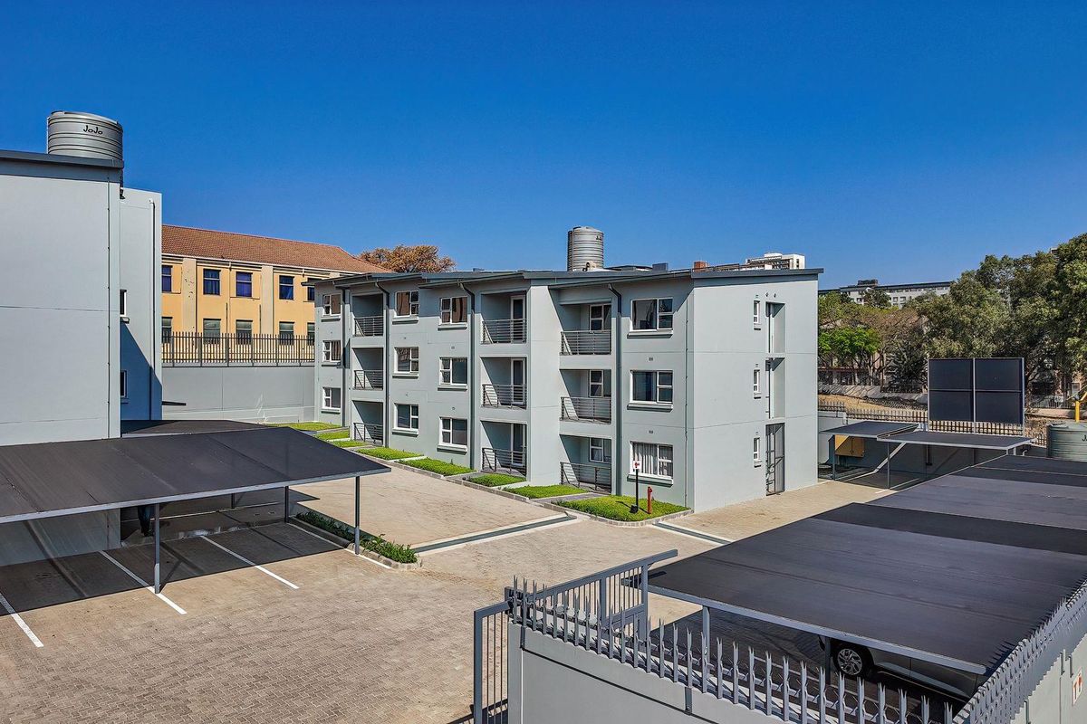 Aerial view of an apartment complex featuring paved parking areas with covered bays, security fencing, and multiple residential units with balconies.