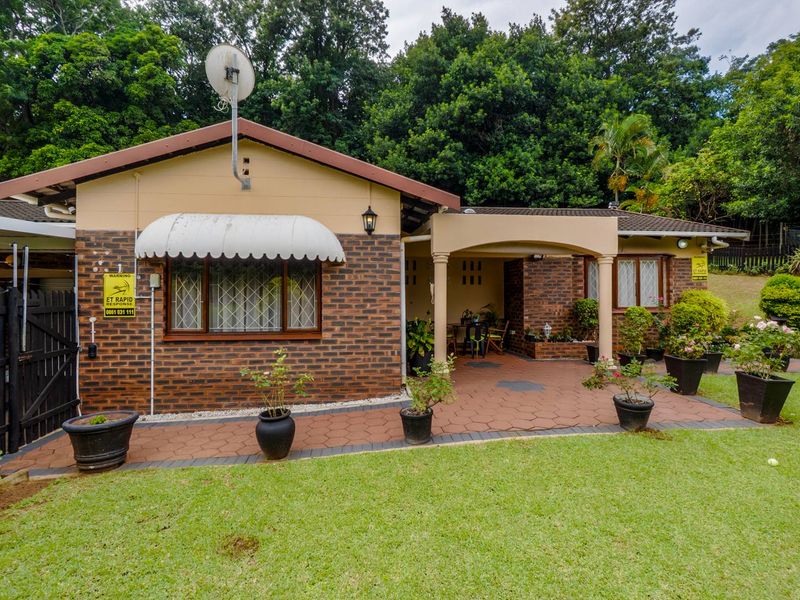 A charming townhouse exterior featuring a brick facade, covered patio area, and a well-maintained garden setting with mature trees in the background.