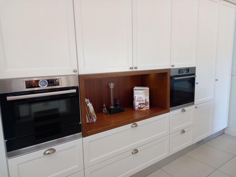 Modern kitchen featuring white shaker-style cabinetry, integrated stainless steel ovens, and a contrasting timber-look open shelving unit.