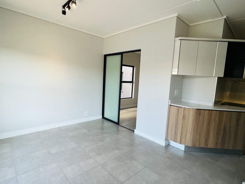 Modern open-plan living area featuring neutral tiled flooring, track lighting, and a compact kitchen with contemporary cabinetry and integrated appliances.