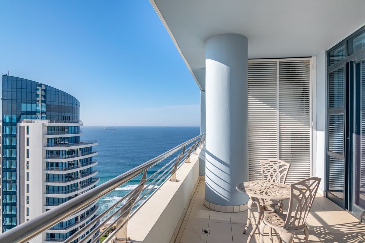 A spacious balcony featuring a stainless steel railing, tiled flooring, and a structural column, offering expansive ocean and city skyline views.