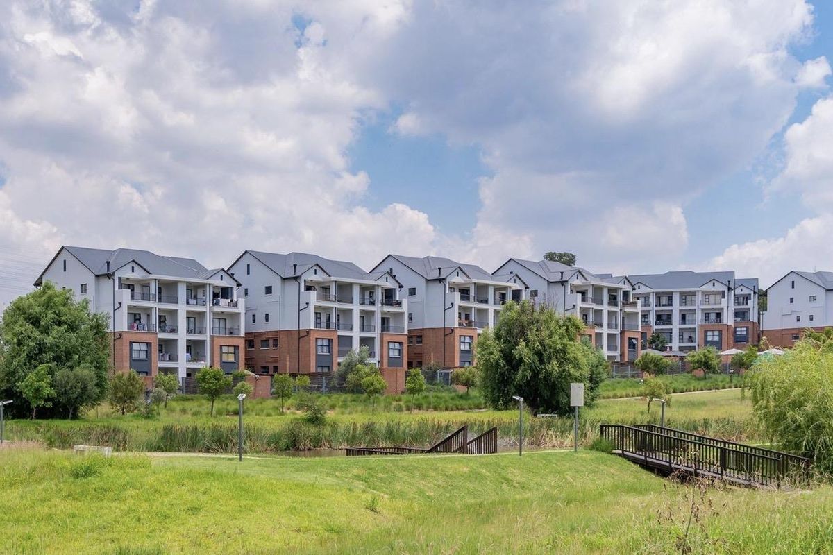 A view of the rear elevation of a modern apartment complex featuring multi-story blocks with balconies, set within a landscaped estate with a pedestrian bridge.