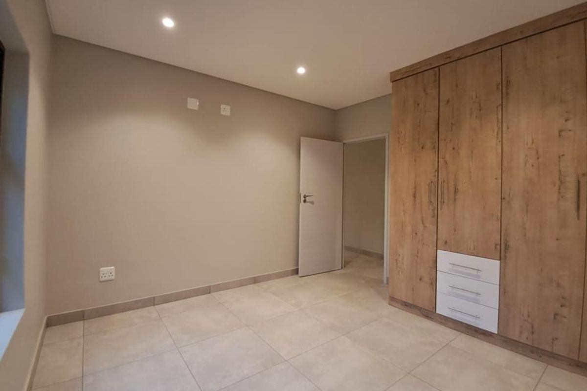 A bright, modern bedroom featuring neutral tiled flooring, recessed ceiling lighting, and built-in wooden cabinetry with integrated drawers.