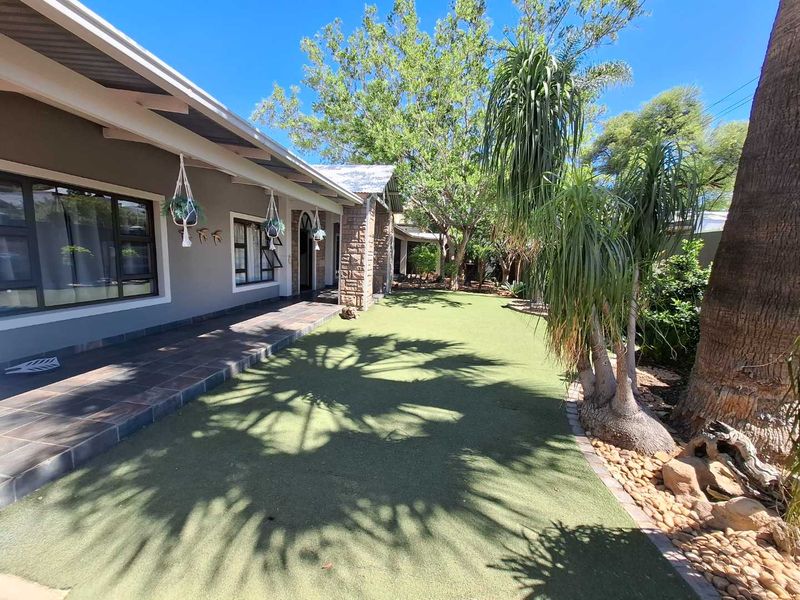 A well-maintained rear garden featuring a tiled patio, stone-clad pillars, and a neat artificial lawn area under mature trees.