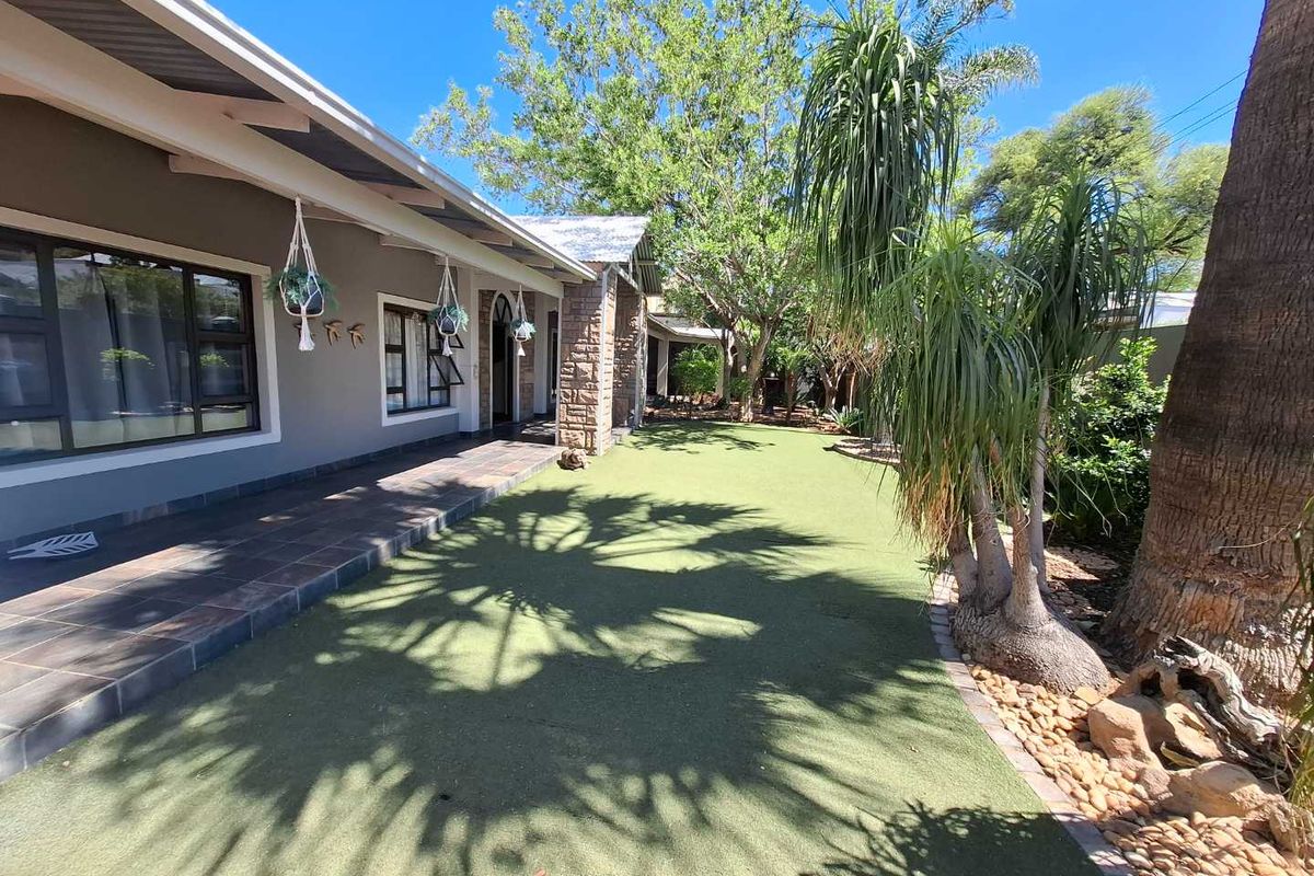 A well-maintained rear garden featuring a tiled patio, stone-clad pillars, and a neat artificial lawn area under mature trees.