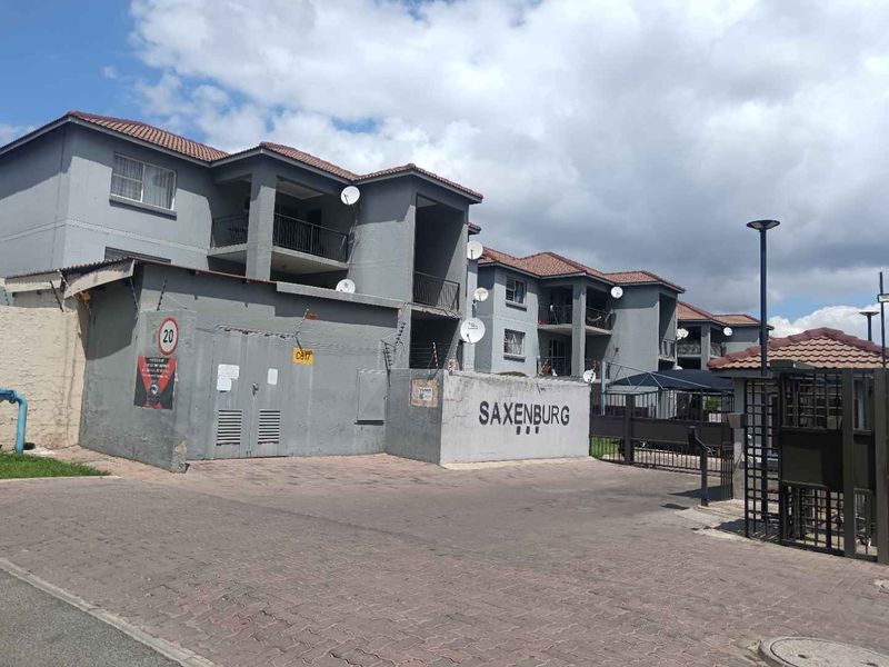 Front view of the Saxenburg apartment complex featuring a paved entrance, security gate, and multi-story residential units with balconies.