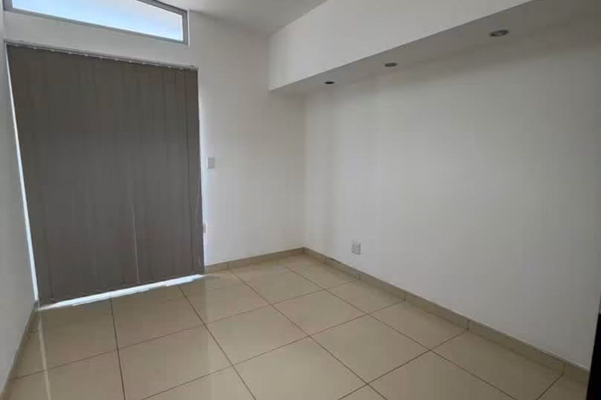 A bright, empty bedroom featuring neutral tiled flooring, recessed ceiling lighting, and a clerestory window with vertical blinds.