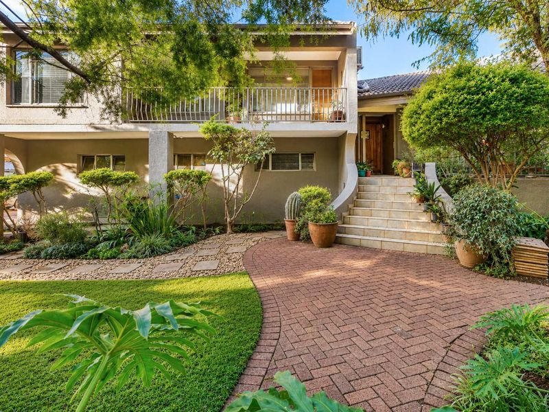 A well-maintained front facade featuring a brick paved entrance, stone staircase, and a balcony overlooking a lush, landscaped garden.
