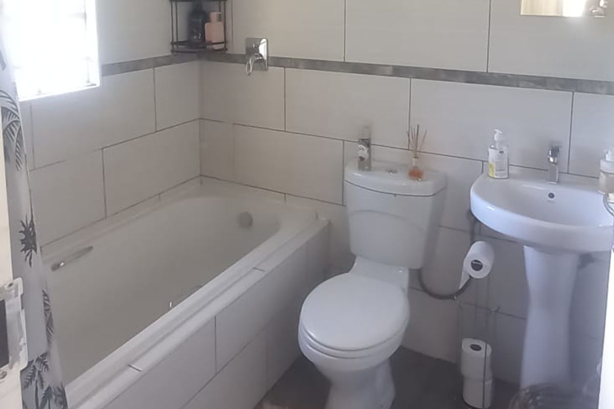 A functional bathroom featuring a built-in bathtub, pedestal washbasin, and dual-flush toilet with neutral wall tiling and grey floor tiles.
