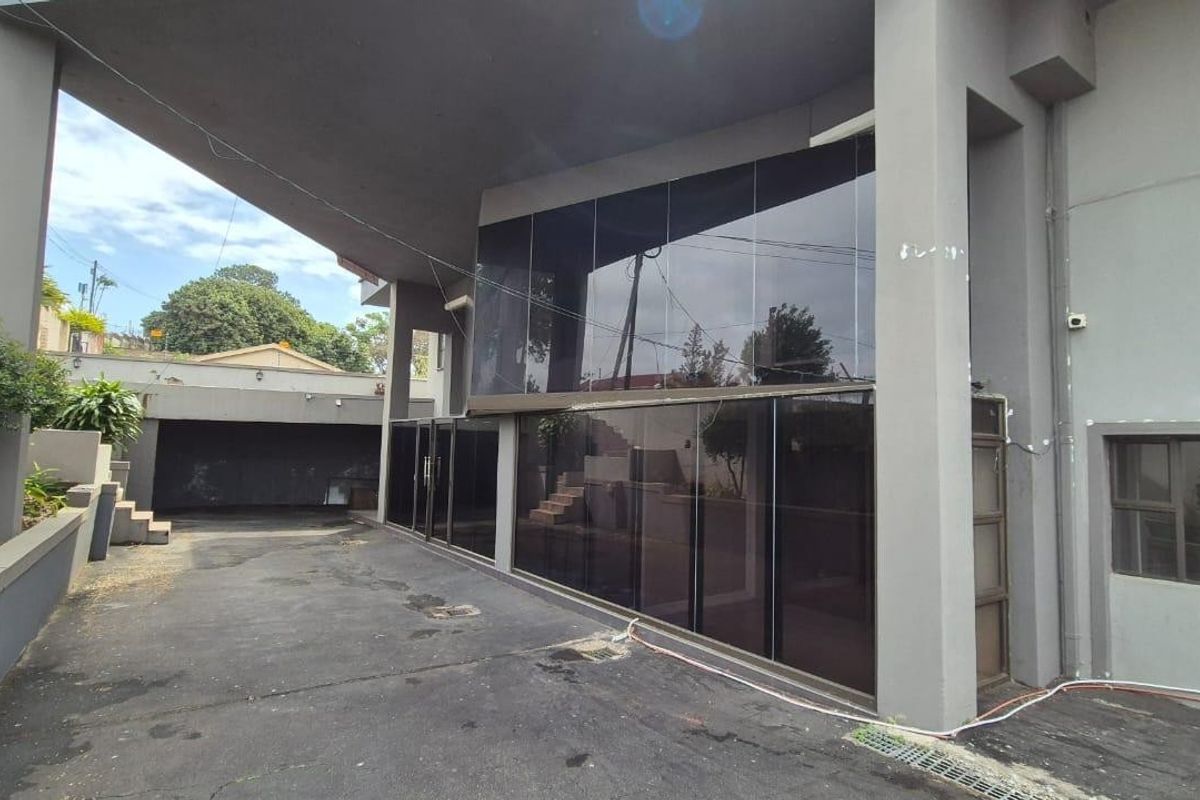 Rear exterior view featuring a large asphalt driveway, covered patio space, and a modern tinted glass facade with concrete structural pillars.
