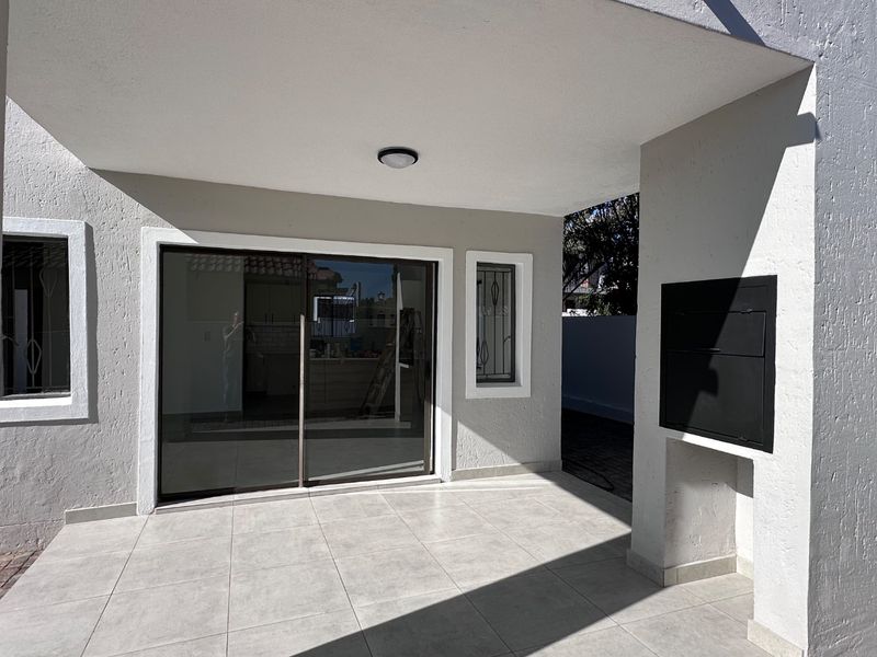 A clean, covered patio area featuring modern tiled flooring, a built-in braai, and sliding glass doors providing access to the interior.