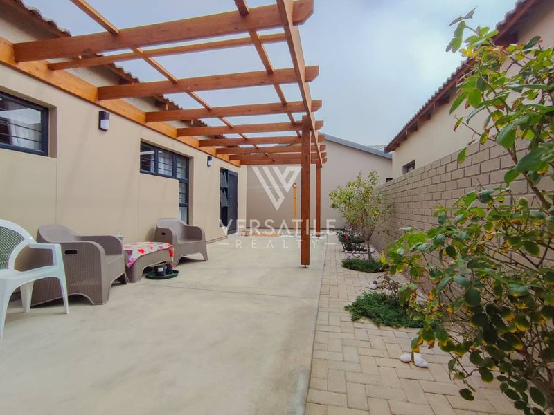 A well-maintained outdoor patio area featuring a timber pergola, concrete flooring, and a paved garden path alongside a brick boundary wall.