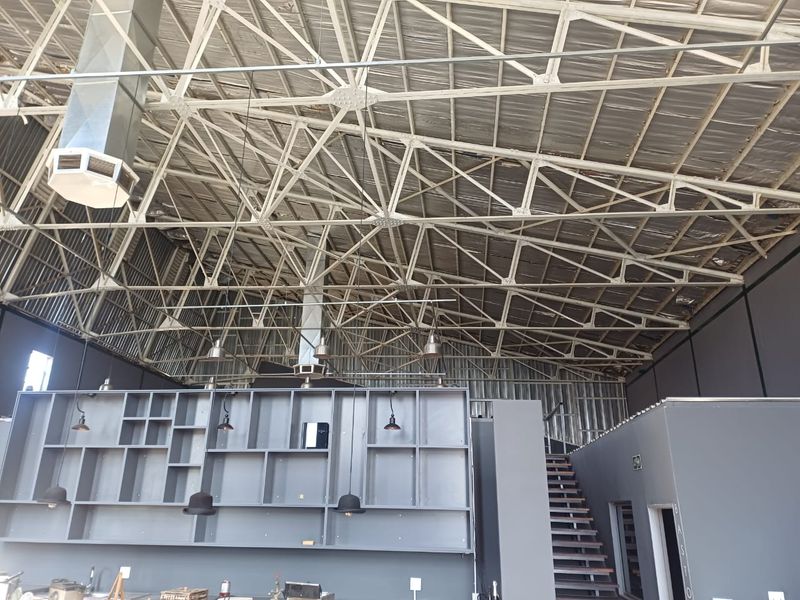 Spacious open-plan industrial-style interior featuring high exposed steel roof trusses, metal roofing, built-in shelving, and a staircase leading to an upper level.