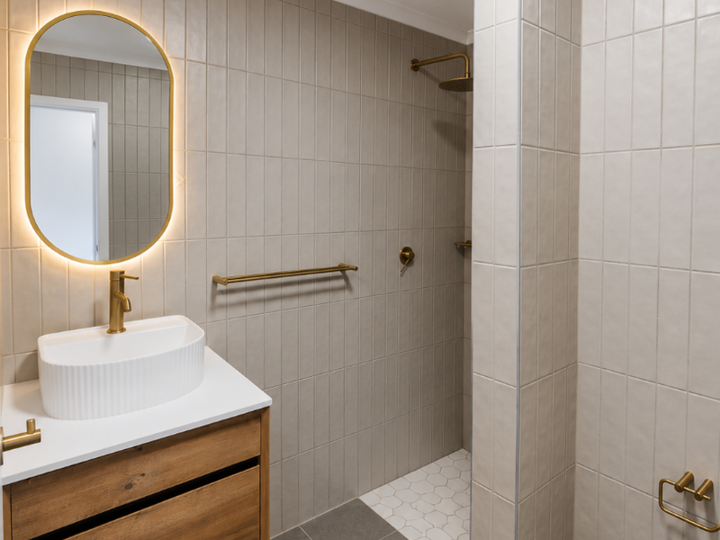 Modern bathroom featuring a floating timber vanity, vessel sink, brass fixtures, backlit mirror, and a walk-in shower with neutral ceramic tiling.