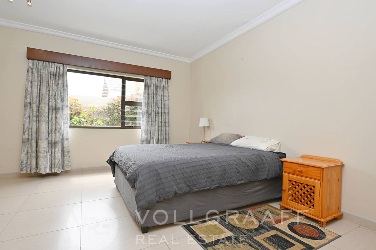 A spacious bedroom featuring neutral tiled flooring, crown molding, and a large window with garden views.